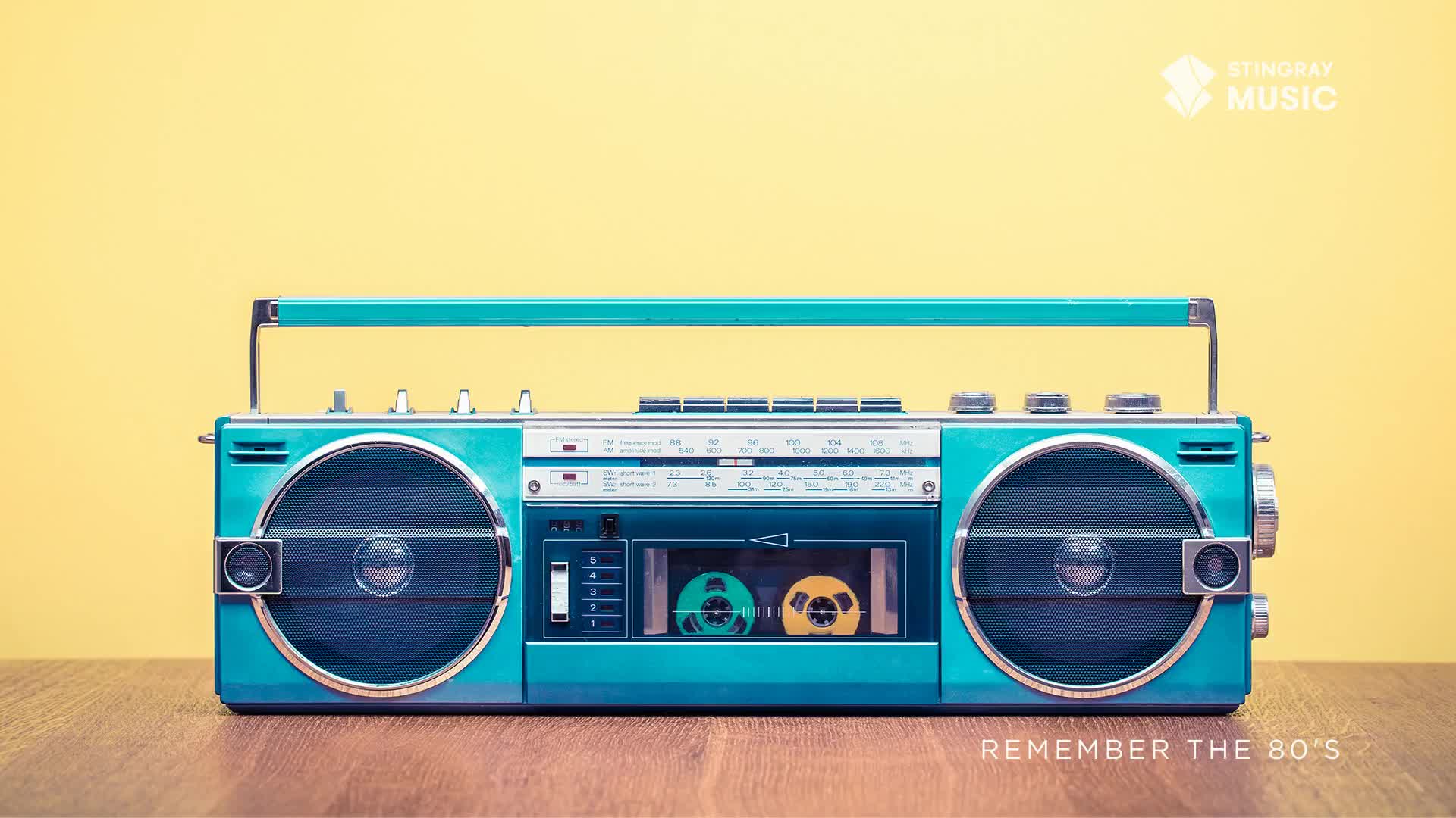 A turquoise boombox, a relic from the Stingray Everything '80s era, sits on a wooden surface against a bright yellow backdrop. Two cassette tapes are visible inside, ready to blast those classic Canadian hits.
