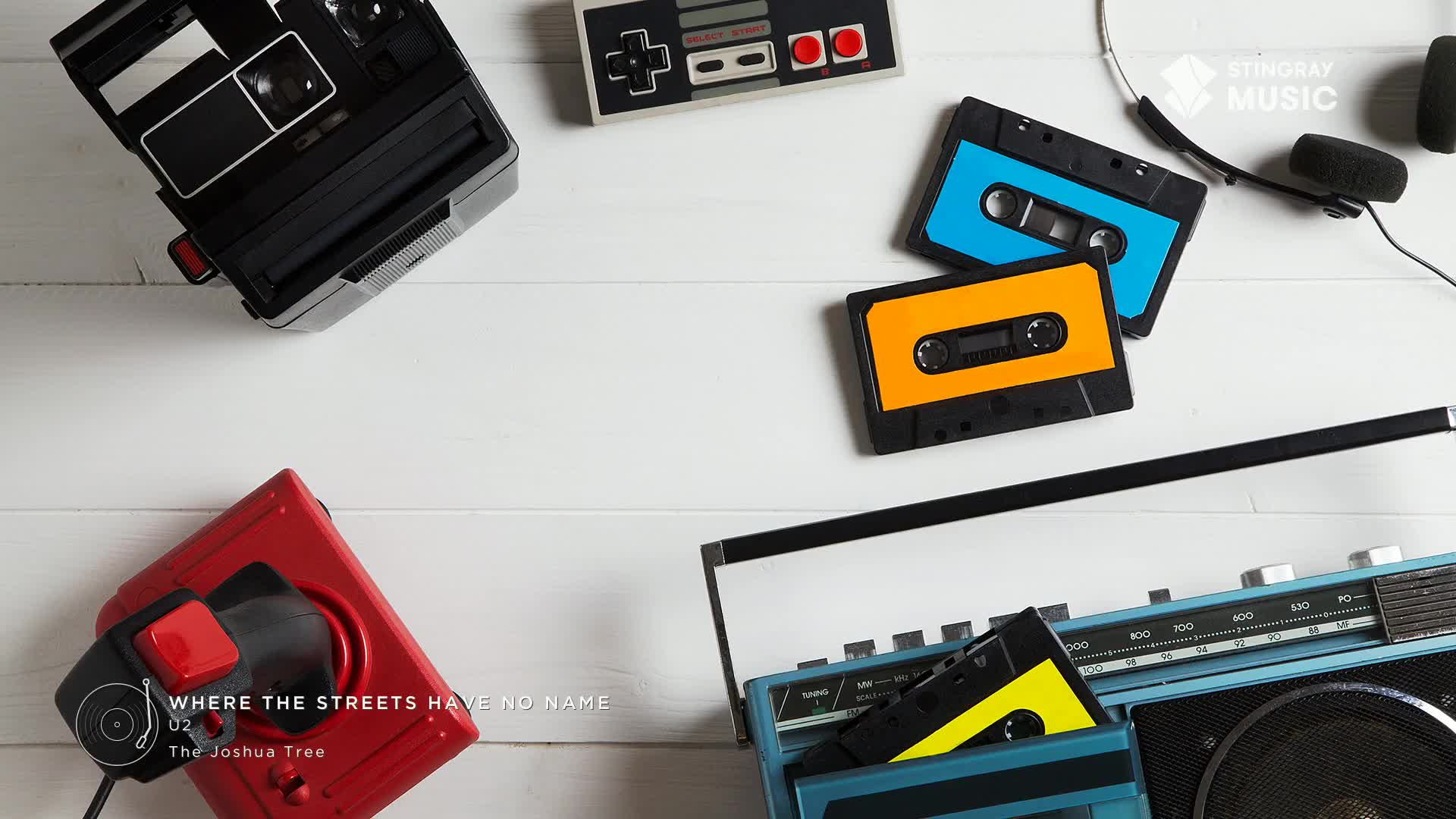 A red joystick and a black camera sit beside a retro boombox, with colorful cassette tapes scattered across the white surface. The Stingray Music logo and the "Where the Streets Have No Name" title card suggest a blast from the past.
