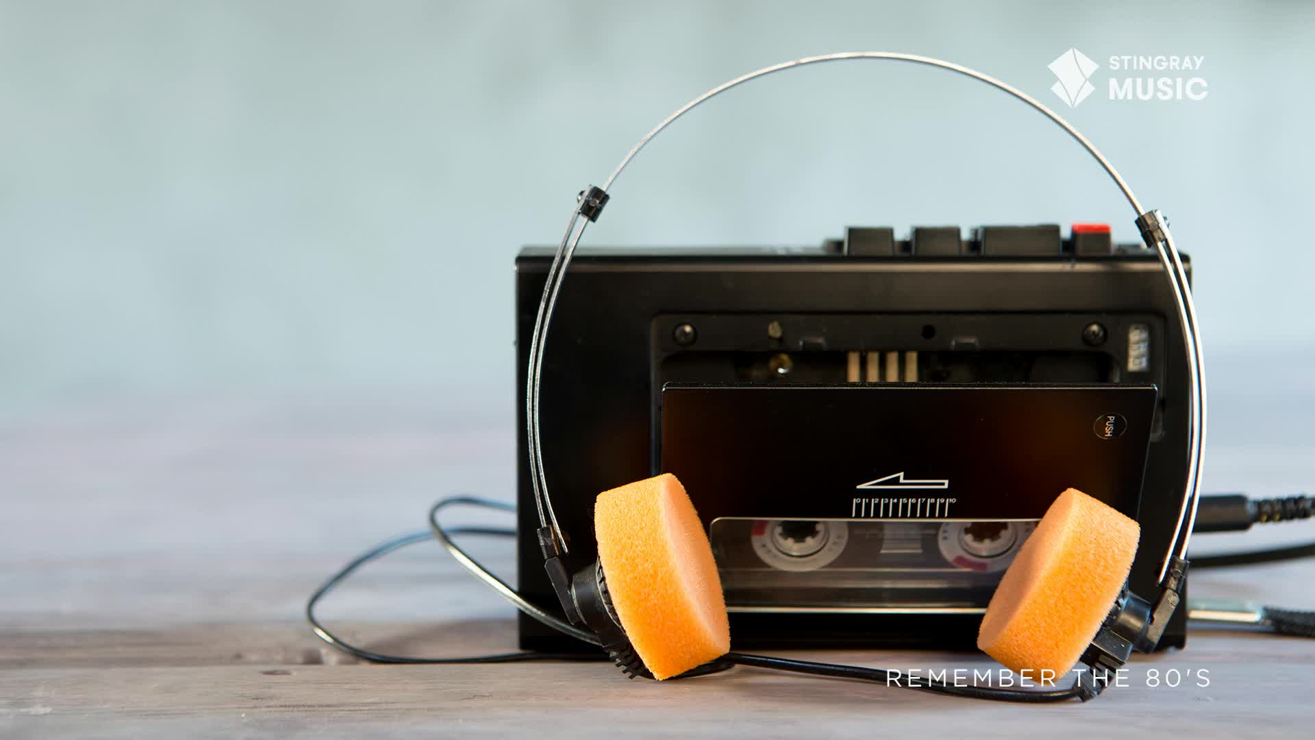 A black cassette player sits on a wooden surface, with the headphones draped over it. The orange foam earpads rest against the player, ready for a blast from Stingray Everything '80s.
