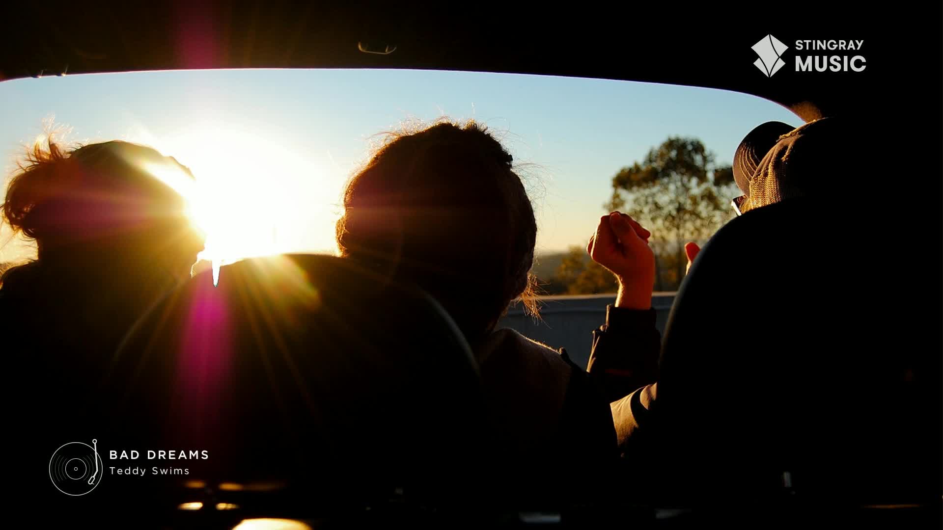 The sun blazes through the car window, silhouetting the heads of the passengers. A hand gestures towards the landscape as "Bad Dreams" by Teddy Swims plays on the Stingray Hit List.
