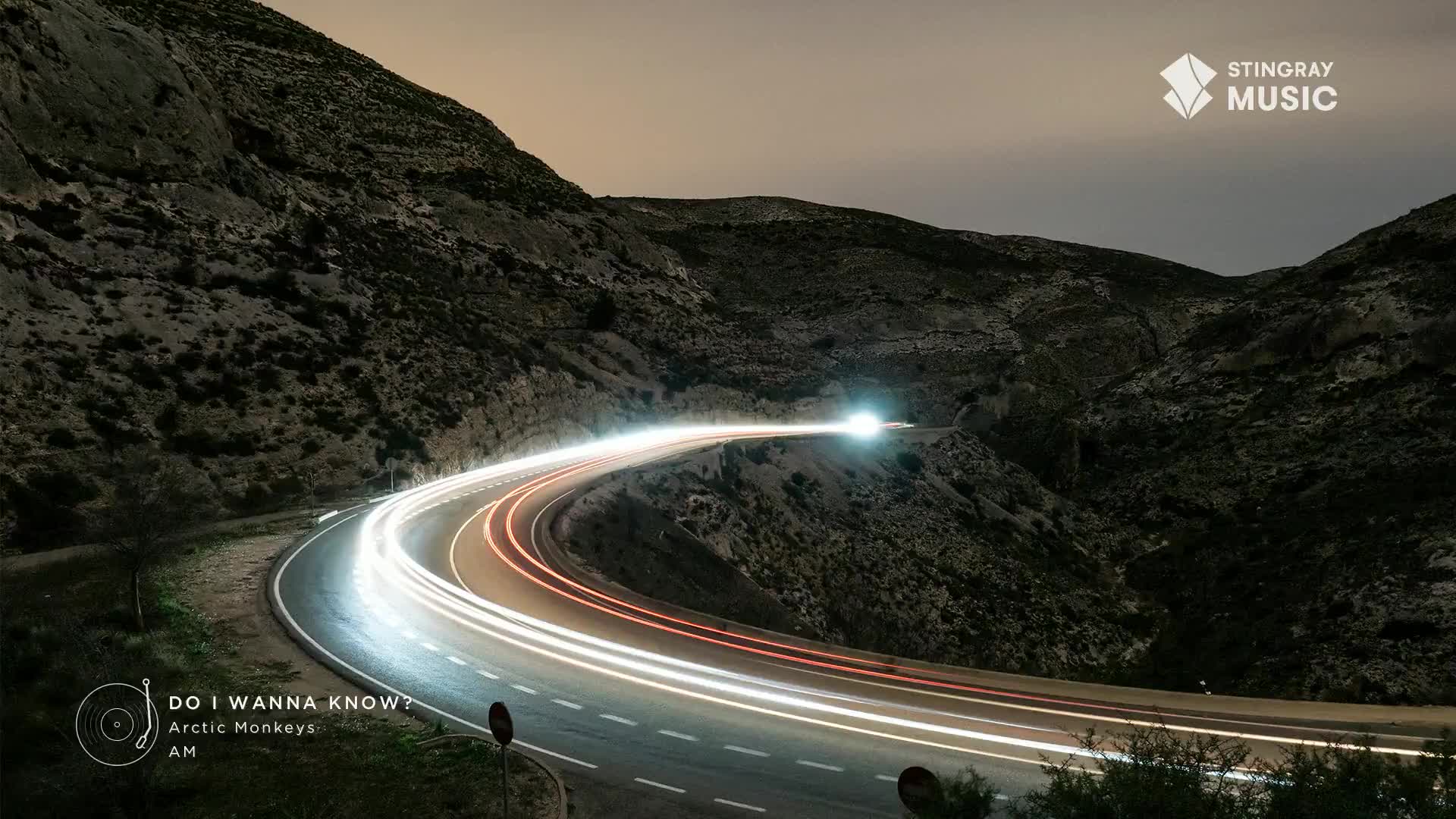 Headlights streak through the winding road, leaving glowing trails of white and red against the dark mountainside. The Stingray Hit List logo sits in the upper right corner, and the song "Do I Wanna Know?" by Arctic Monkeys plays in Canada.

