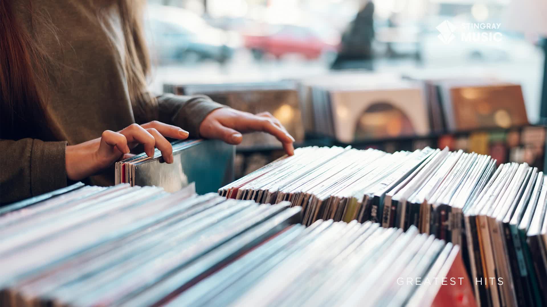 A person's hands are carefully sifting through a bin of records, perhaps searching for a Stingray Holiday Hits album. The spines of the records are visible, and the light catches the edges of the covers.
