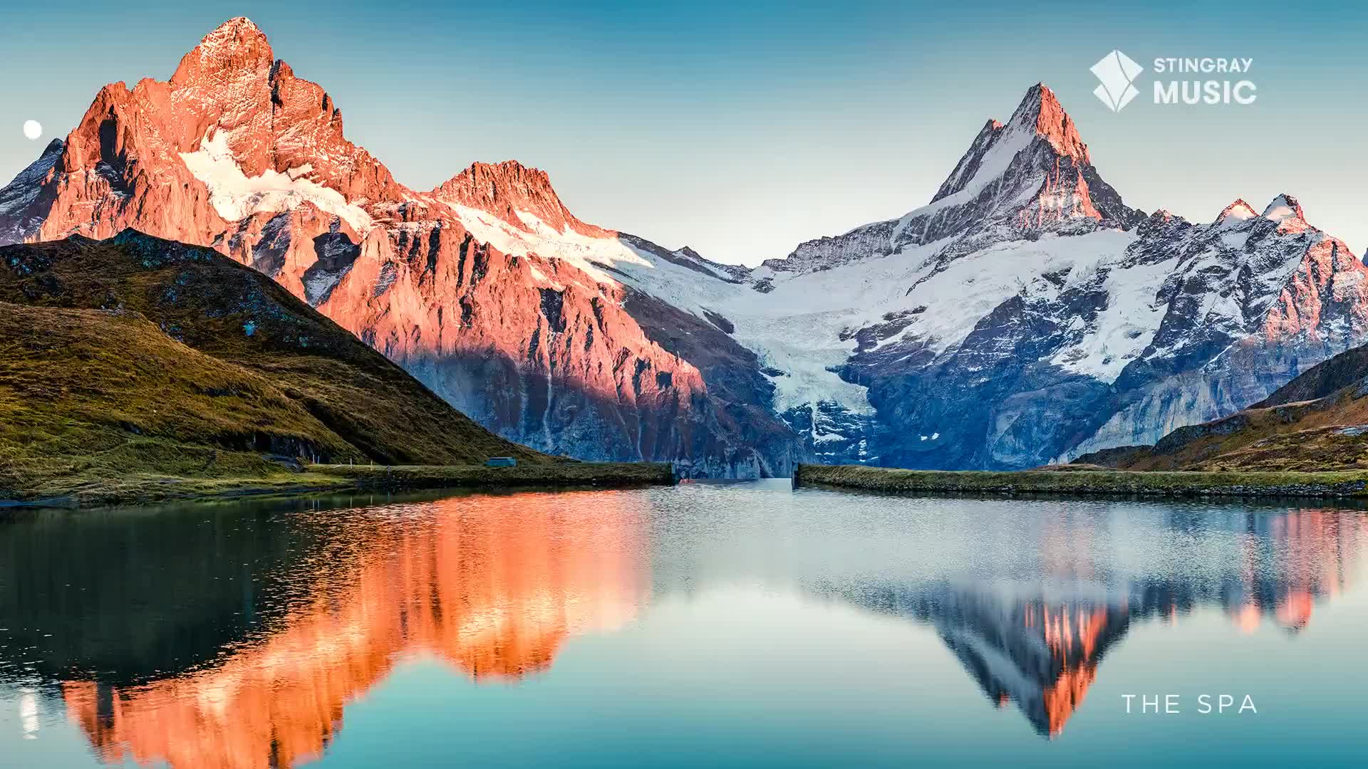 The sun hits the jagged peaks, casting a warm, orange glow across the mountainside. A calm lake perfectly mirrors the scene, reflecting the snow-capped summits and the clear, blue sky.