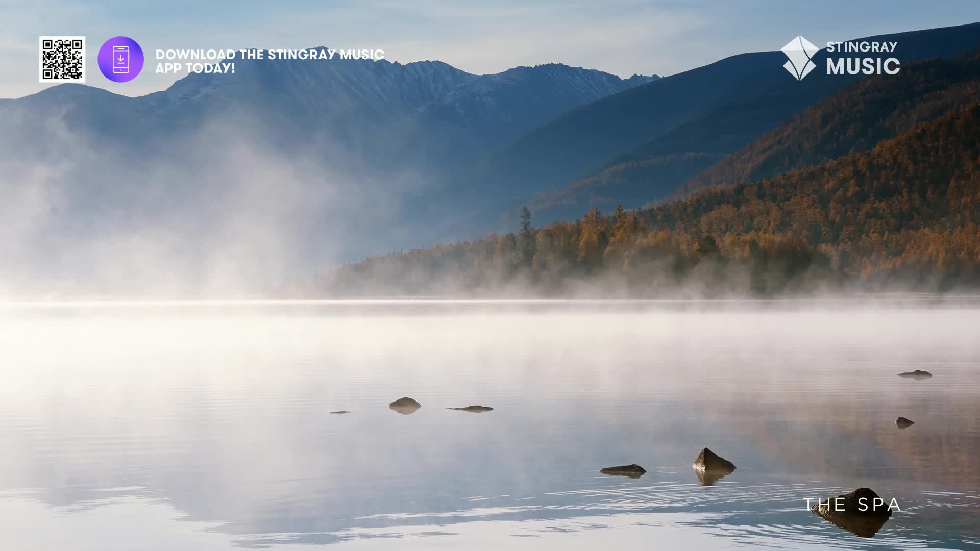 Mist rises from the calm, glassy surface of a Canadian lake, reflecting the rugged, tree-lined mountains beyond. A few dark rocks break the water's surface, hinting at the tranquil depths of The Spa.