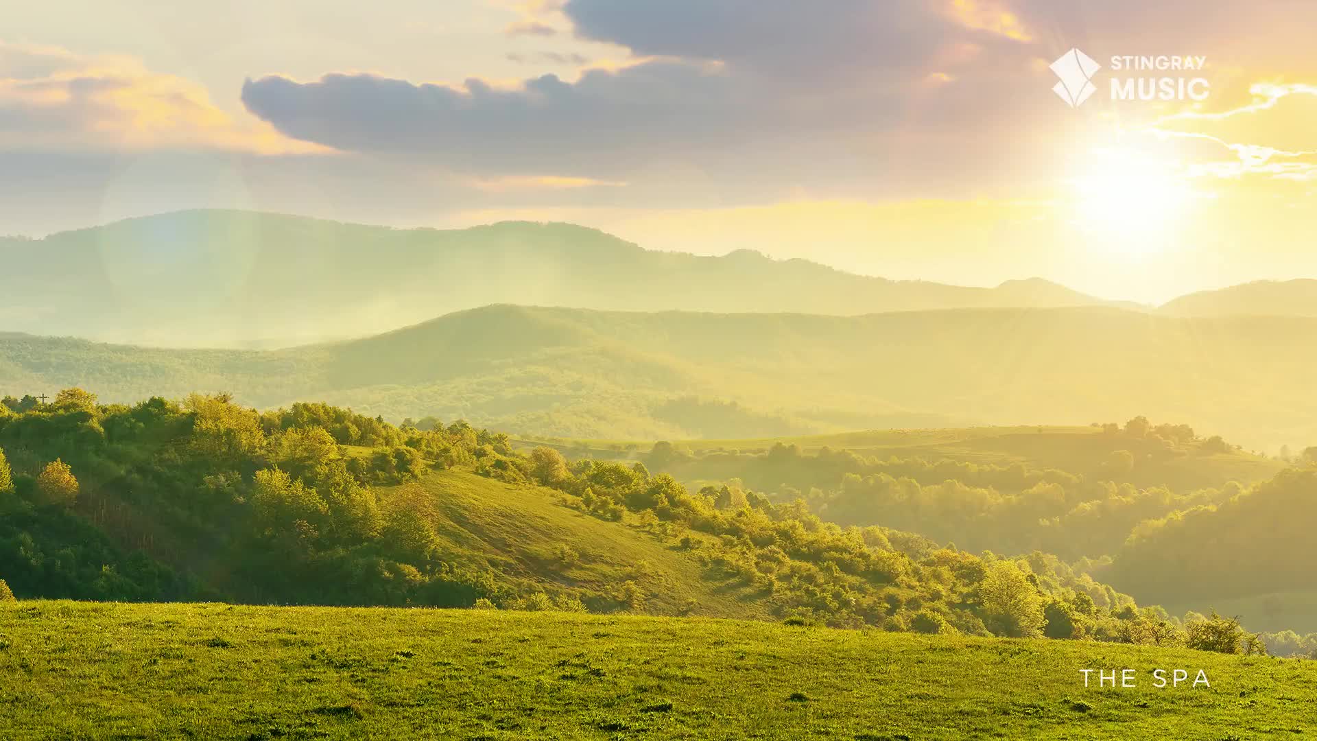 The sun breaks through the clouds, casting a golden haze over rolling Canadian hills. Lush green fields and scattered trees stretch towards a distant, misty mountain range.