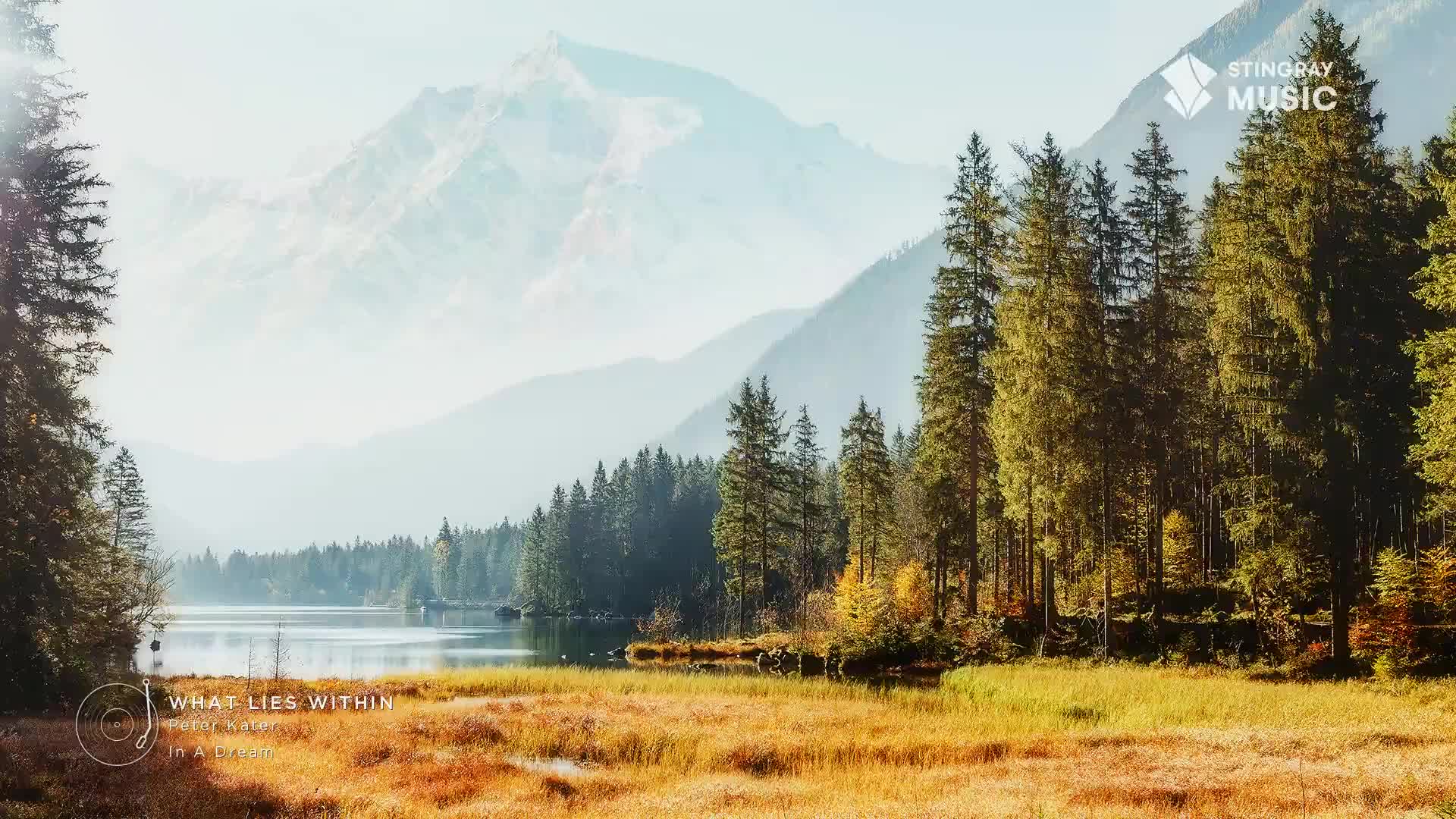 The sun warms the tall pines lining the lake in this Canadian landscape. Distant mountains fade into a hazy blue sky.