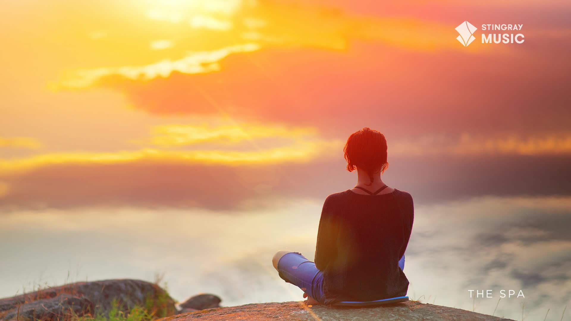The sky glows with a warm, orange light as clouds drift by. A person sits cross-legged on a rock, facing the sunset.