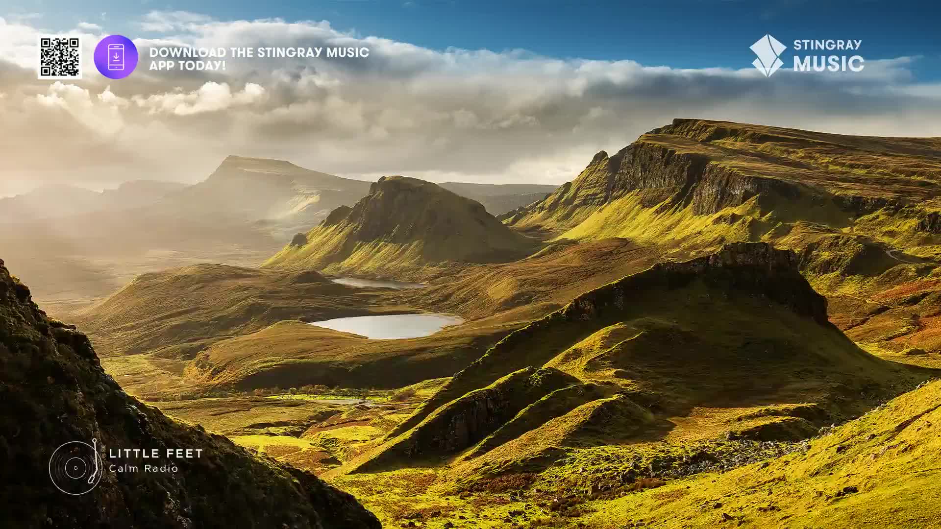 The sun casts long shadows across the rugged, green hills of Canada. A calm lake sits nestled in the valley below, reflecting the dramatic sky.