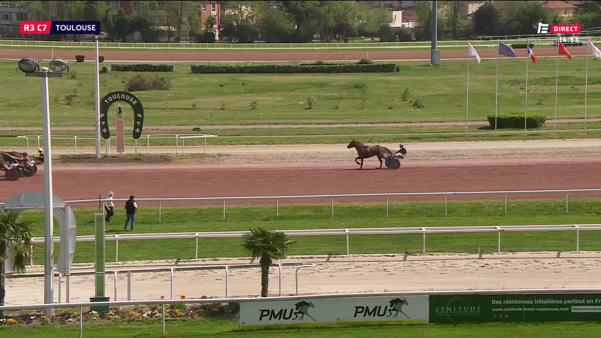 A horse pulls a sulky down the dirt track. Flags wave in the background near the finish line.