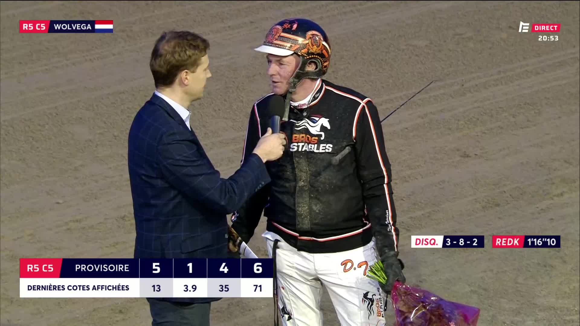 A man in a racing helmet and silks is being interviewed by a reporter. He holds a bouquet of flowers, the colors of the French flag visible on his helmet.