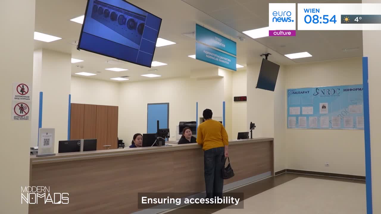 A person in a yellow sweater turns to speak with someone behind a counter. The scene unfolds in a modern office space, likely a public service building, with screens displaying information.