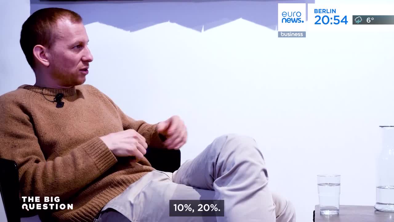 A man in a tan sweater gestures with his hands as he speaks, his light-colored pants creased. A screen in the background displays "euronews. berlin 20:54" and "business."