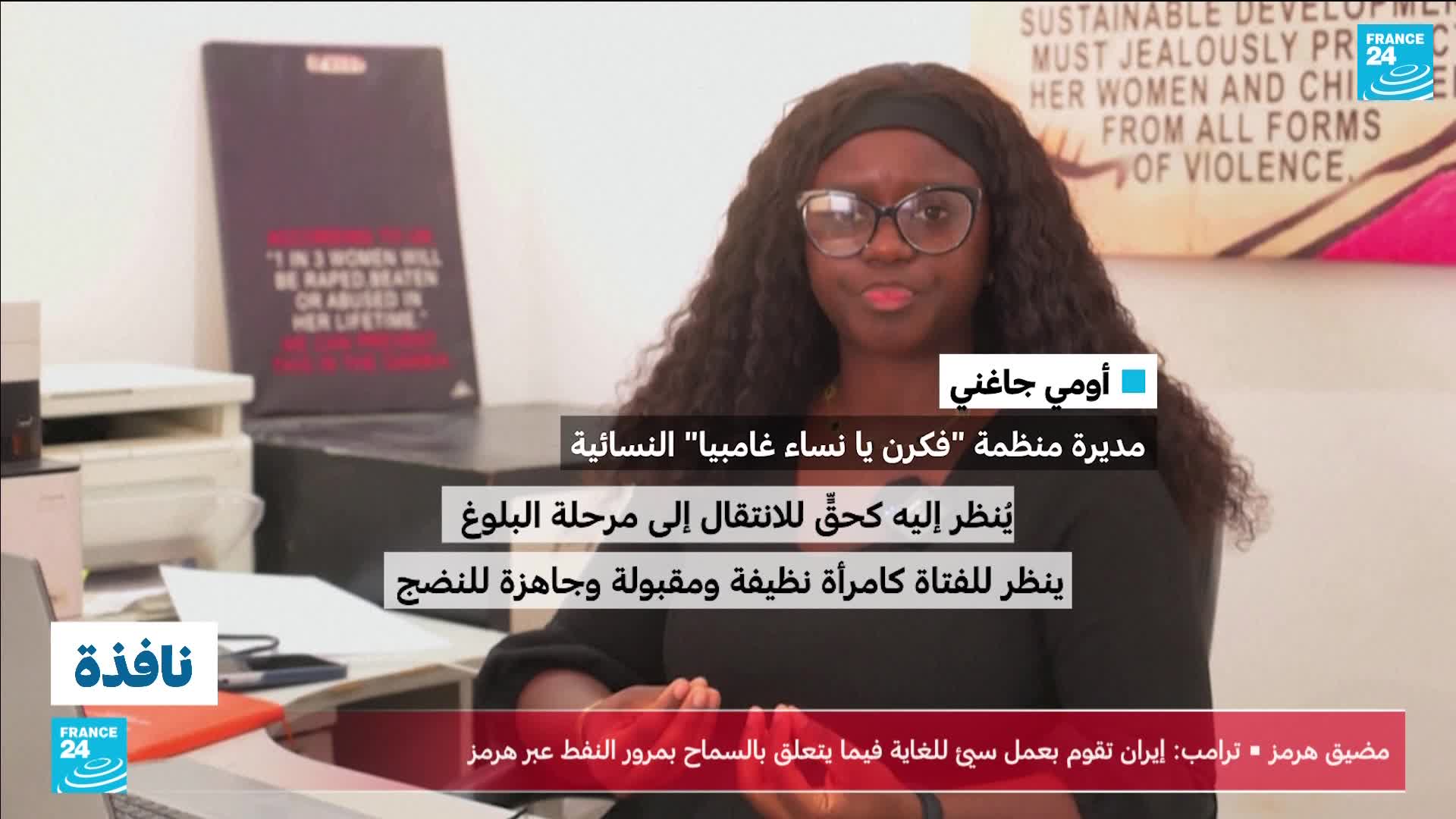 A woman with glasses and long, dark hair speaks directly to the camera. Behind her, a poster displays a stark message about violence against women.