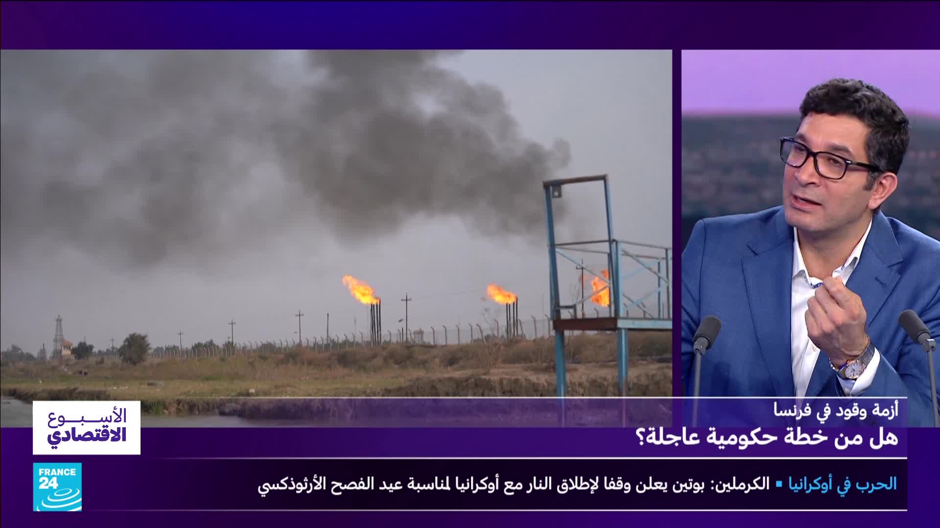Flames erupt from tall stacks, sending thick black smoke into the overcast sky. A man in a blue suit gestures with his hands while speaking, his glasses reflecting the studio lights.