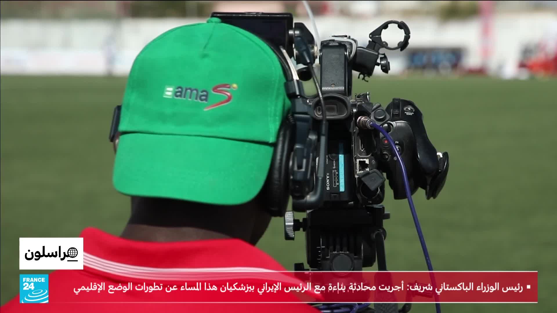 A cameraman, wearing a bright green cap, steadies a professional video camera on a tripod. The camera is pointed towards a green field where figures in red are visible in the distance.