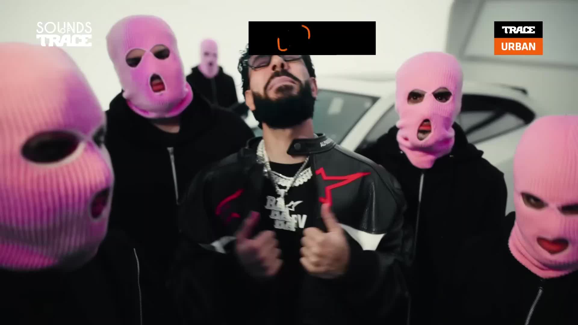 A man in a leather jacket stands surrounded by people in pink ski masks. The Trace Urban logo appears on the right side of the screen.
