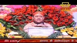 A man in a turban speaks from behind a podium, a backdrop of vibrant red and yellow flowers framing him. The broadcast is from India, and the Aastha Bhajan logo is visible.
