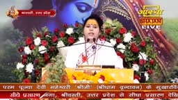 A woman in a white sari speaks into microphones, her face lit by studio lights. Behind her, a floral arrangement of red and white roses frames the stage, and the Aastha Bhajan logo is visible.
