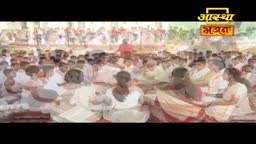 People in white clothing sit closely together, facing a central figure in red, likely engaged in an Aastha Bhajan gathering. The logo in the corner confirms the context of India and the religious event.
