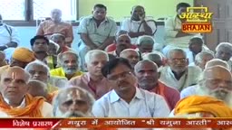 A crowd of men, many with white beards and saffron-colored head coverings, sit attentively. The Aastha Bhajan logo is visible, and the scene is likely from a religious gathering in India.
