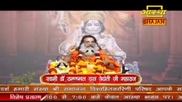 A man with a long white beard and orange robes sits before a large image of a deity, his hands raised. The Aastha Bhajan logo is visible in the upper right corner.