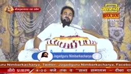 A man in a white kurta speaks into a microphone, his face framed by a dark beard. Before him, a large, circular offering sits on a stand, adorned with alternating red and white striped fabric. The Aastha Bhajan logo appears in the upper right corner.