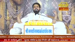 A man in white sits behind a podium, speaking into a microphone. He gestures with his hands as he addresses the audience, his face illuminated by the stage lights.