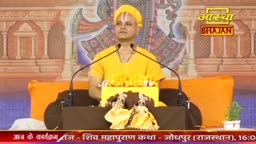 A man in a saffron robe and turban speaks into a microphone, his hands resting on a yellow podium adorned with marigolds. Behind him, a colorful backdrop depicts temple architecture, with the Aastha Bhajan logo visible in the upper right corner.