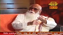 An elderly man with a long white beard, wearing glasses and a white kurta, gestures with his hands as he speaks. The Aastha Bhajan logo is visible in the background.