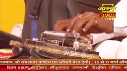 A man's hands, adorned with rings, are carefully adjusting a miniature, ornate temple. The Aastha Bhajan logo is visible in the background.