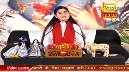 A woman in a white outfit and red scarf speaks into microphones, her long dark hair falling around her. Beside her, on a raised platform, a small, patterned animal figurine stands to the right of a wooden box adorned with flowers.