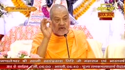 An elder in an orange robe speaks into a microphone, his right hand raised with his thumb and index finger touching. Behind him, a banner displays "Aastha Bhajan" in Hindi.