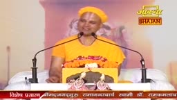 A man in an orange robe and yellow cap speaks into two microphones. He stands behind a podium adorned with marigolds and a small statue.