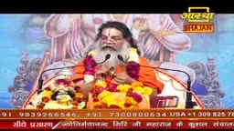 A man with a long white beard, wearing orange robes, speaks into two microphones. He sits behind a large arrangement of yellow and orange flowers.