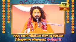 A man in an orange robe speaks into a microphone, his long hair framing his face. The backdrop is a warm orange, with strings of marigold garlands adorning the sides.