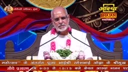 A man with a white beard and glasses speaks into a microphone, wearing a white kurta and a garland of pink flowers. The Aastha Bhajan logo is visible in the background.