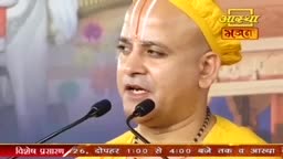 A man in a yellow cap and saffron robe speaks into two microphones, his forehead marked with a white and red tilak. The Aastha Bhajan logo appears in the upper right corner.
