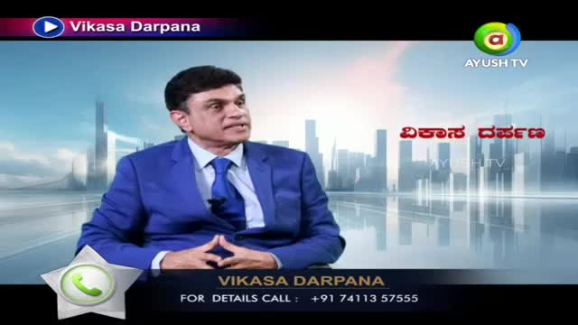 A man in a sharp blue suit speaks on Ayush TV, his hands clasped before him. The program title, "Vikasa Darpana," appears in Kannada script behind him.