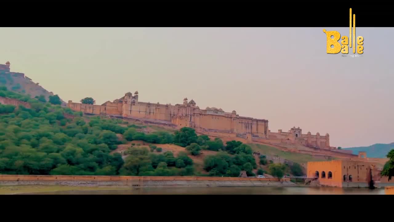 The Amber Fort stretches across the hillside, its sandstone walls glowing in the soft light. A calm lake reflects the ancient architecture, with a small, arched building nestled by its edge.
