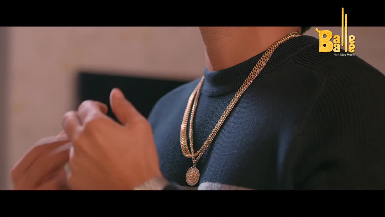 A man's hands adjust a thick gold chain draped over his dark sweater. The Balle Balle logo appears in the corner, suggesting a Punjabi music video.