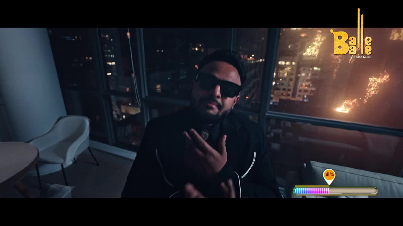 A man in sunglasses gestures with his hands, his face illuminated by the city lights outside the window. The "Balle Balle" logo hovers above the urban glow.