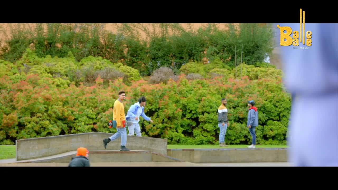 A young man in a yellow shirt walks with a skateboard, while another in blue gestures. Two others stand watching in the background.