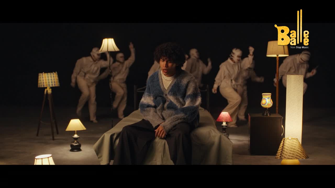 A young man sits on a bed, surrounded by a troupe of dancers in beige jumpsuits, their movements a stark contrast to his stillness. The Balle Balle logo gleams in the corner, a reminder of the vibrant Indian music scene.