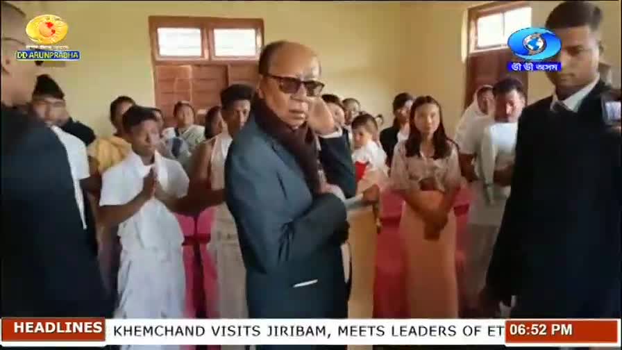 A man in a dark suit and scarf, wearing sunglasses, speaks on his phone as a crowd gathers around him. A young man in traditional white attire folds his hands in greeting.