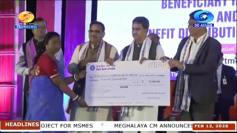 A woman in a blue sari and red blouse accepts a large check from two men in traditional Indian attire. The State Bank of India logo is visible on the check, and the DD Arun Prabha logo appears on a screen behind them.