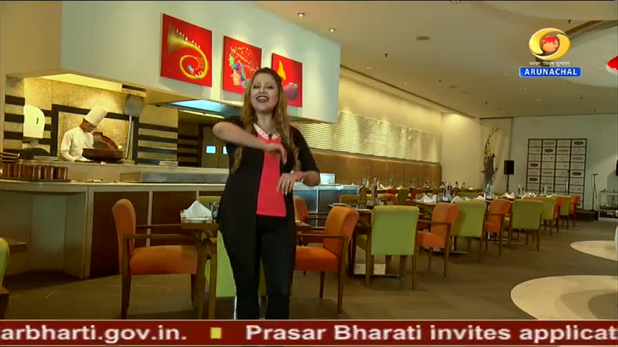 A woman in a black and red outfit gestures enthusiastically in a restaurant. Behind her, a chef in a white uniform works near a pizza oven.