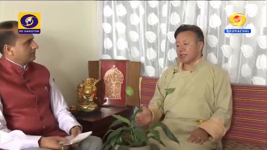 A man in a maroon vest and white shirt speaks with a person in a traditional tan robe, both seated on a striped couch. The DD Arun Prabha logo, indicating a broadcast from India, appears in the upper right corner.