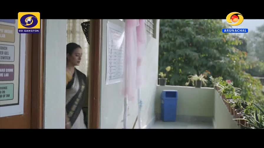 A woman in a sari pauses at a doorway, her gaze directed inward. Outside, potted plants line a balcony overlooking lush green foliage.