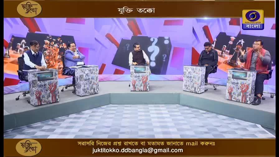 Five men are seated on chairs behind podiums on a stage, facing a camera. The backdrop behind them is a colorful, abstract design with question marks. The logo for DD Bangla is visible in the upper right corner.