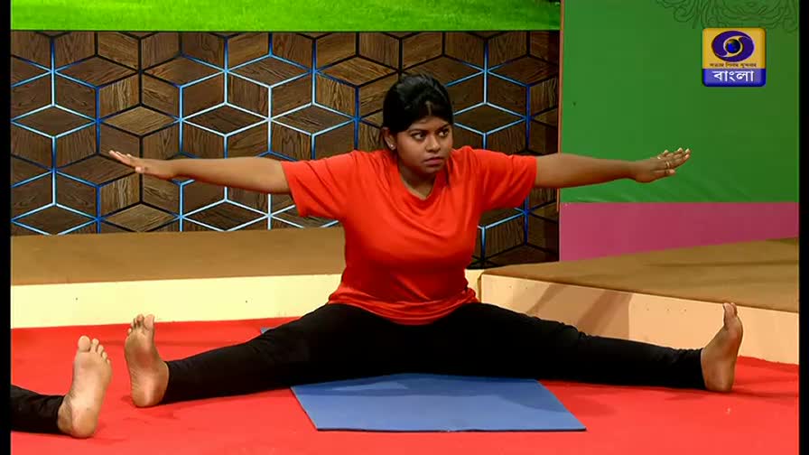 A woman in a bright orange shirt and black leggings holds a wide straddle pose on a blue mat. Her arms extend outwards, parallel to the floor, as she focuses intently on her movements.